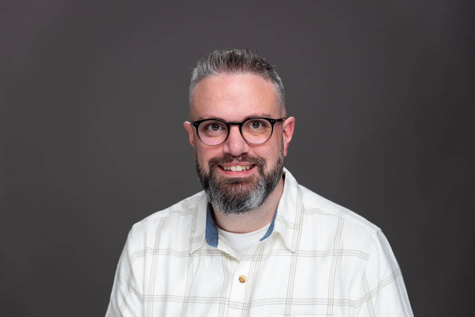 A headshot of Zach McCurdy, a man with glasses and a beard, smiling in a white plaid shirt against a dark grey studio background.